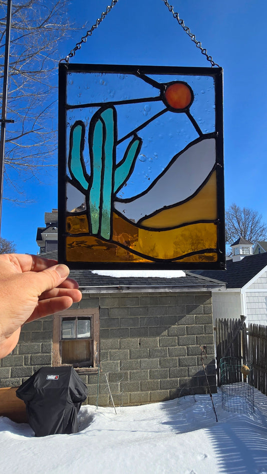 Staine glass panel featuring a bold green cactus, warm orange sun, and layered desert landscape in amber and sandâ€‘toned glass, framed in dark solder lines and approximately 8.5 by 11 inches.