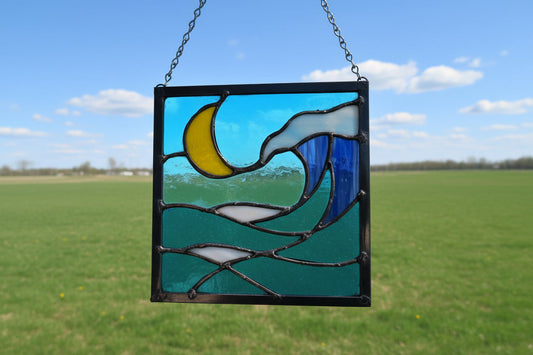 Stained glass panel featuring a yellow crescent moon above layered blue and white ocean waves with a sense of crashing surf, framed in dark solder and approximately 9 by 9 inches.